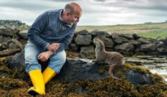 A man in a gray sweater and yellow boots sits on a rocky shore, looking at an otter. The background features cloudy skies and grassy hills.