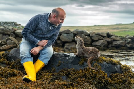 A man in a gray sweater and yellow boots sits on a rocky shore, looking at an otter. The background features cloudy skies and grassy hills.