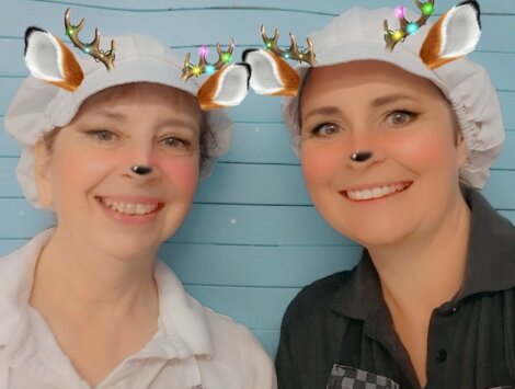 Two people wearing white caps with a deer filter, including antlers and ears, on their heads. They are smiling in front of a blue background.
