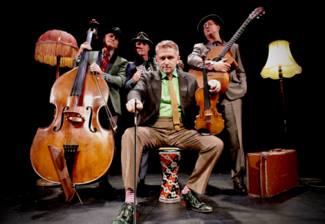 Four musicians in suits pose with instruments, including a double bass, violin, and guitar, on a stage with two floor lamps and a suitcase. The man in front sits on a drum, holding a cane.