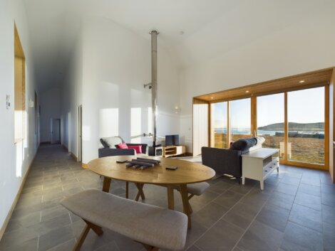Modern living room with a wooden round table, benches, a sofa, tall windows, and a fireplace. Natural light illuminates the space, highlighting the outdoor view.