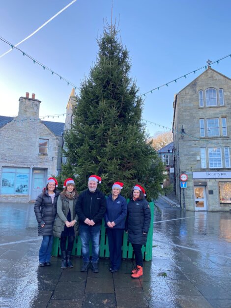 Five people wearing Santa hats stand in front of a large outdoor Christmas tree in a town square with stone buildings.