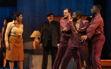 Actors on stage in a dramatic scene; a woman confronts three men in matching maroon outfits, while another man in the background looks on.