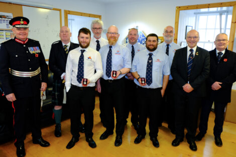 A group of ten men, some in uniforms and others in suits, are standing indoors. Four men in blue shirts are holding medals.