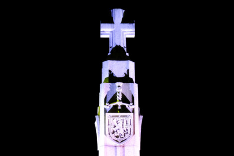 Illuminated stone monument featuring a cross at the top with a shield and figure underneath, set against a dark background.
