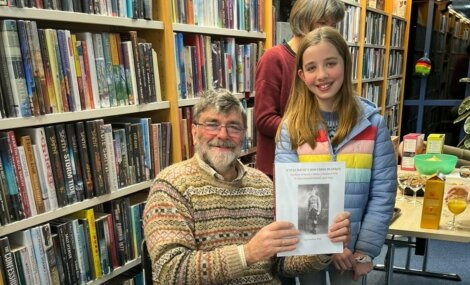 An older man seated in a library holds a book, smiling beside a young girl in a colorful jacket. Shelves filled with books are in the background.