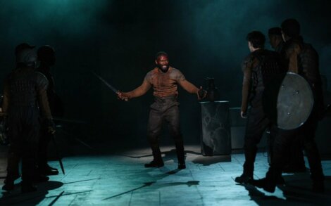 A man holding a sword stands on a dimly lit stage, surrounded by people in dark clothing and armor.