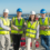 Four people in high-visibility vests and hard hats stand on a balcony with a coastal industrial background.