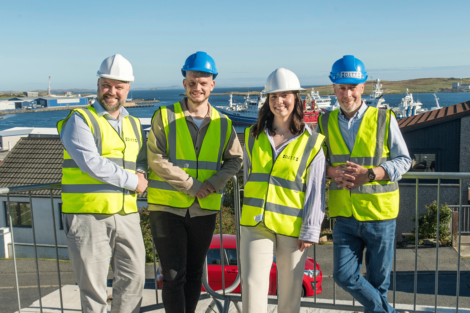Four people in high-visibility vests and hard hats stand on a balcony with a coastal industrial background.