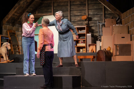 Three people on a stage with boxed props and a hobby horse; one points assertively while another stands between them in a scene.