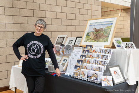 Woman smiling beside a table displaying various art pieces, including framed illustrations and greeting cards.