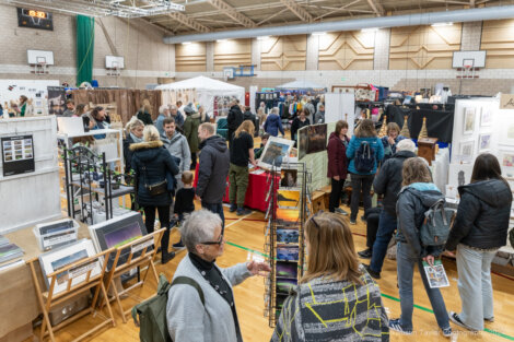 People browse various stalls at an indoor market event held in a large gymnasium.