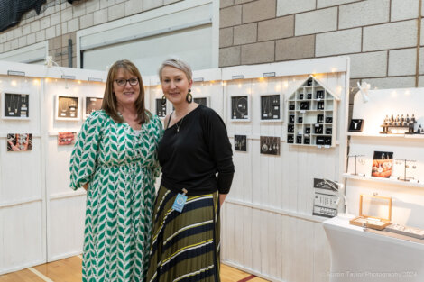 Two women stand smiling in front of a display of jewelry at an indoor exhibition.