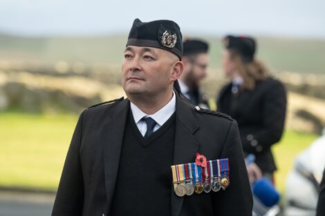 A man in uniform with a black beret and a poppy pin, wearing multiple military medals, stands outdoors.