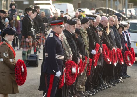 People in uniform and others stand in a line holding wreaths during a remembrance ceremony. A bagpiper is visible in the background.