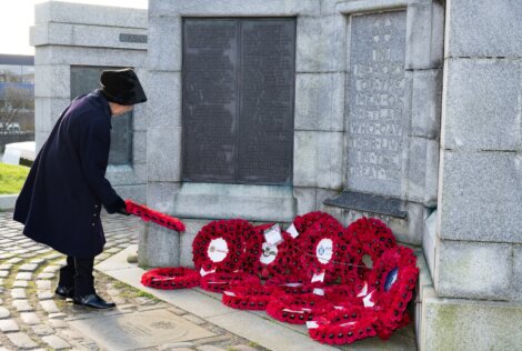 A person places a red poppy wreath at a stone memorial surrounded by other red wreaths.