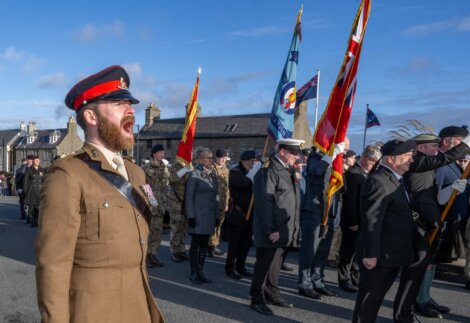 A soldier in uniform shouts commands during a parade with a group of people holding flags in the background.