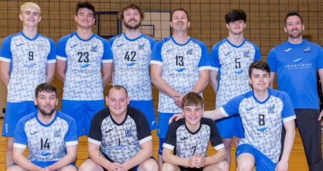 A volleyball team poses in blue and white uniforms in a gym. Ten individuals stand and kneel, smiling at the camera.