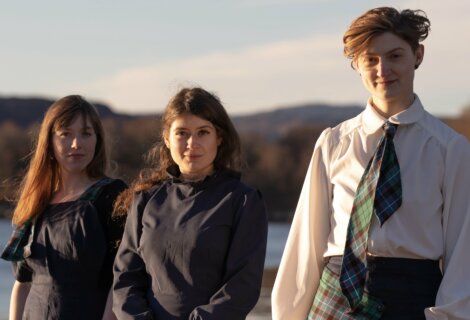 Three people stand outdoors in front of a scenic backdrop, wearing dark outfits. One person wears a tie, and the landscape features hills and water under a clear sky.