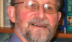 An older man with glasses and a beard, wearing a blue shirt and patterned tie, stands in front of a bookshelf filled with books.