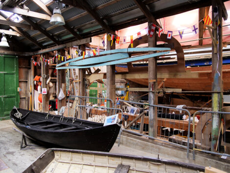 Wooden workshop interior with boats in progress, colorful flags above, and tools scattered around. A black boat is in the foreground, and a blue canoe is suspended from the rafters.