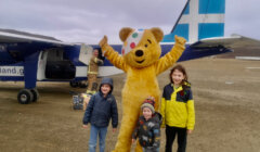 Three children pose with a person in a bear costume in front of a small airplane on an overcast day.