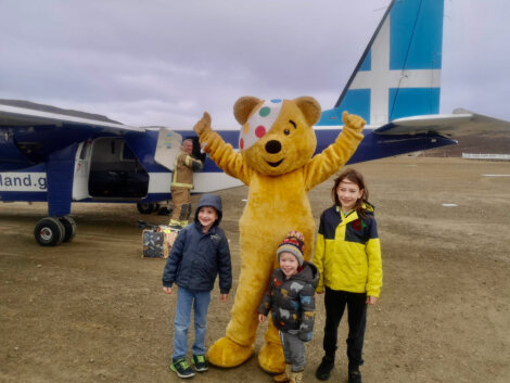 Three children pose with a person in a bear costume in front of a small airplane on an overcast day.