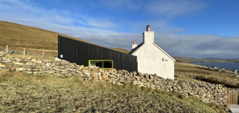 Modern black and white house on a hillside with a stone wall, overlooking a bay under a partly cloudy sky.