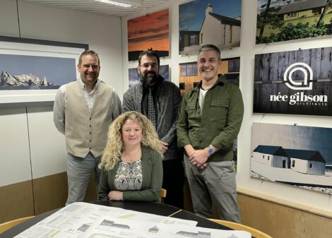 Four people stand in an architect's office, one seated at a table with large paper plans. Walls display various architectural designs and photographs.