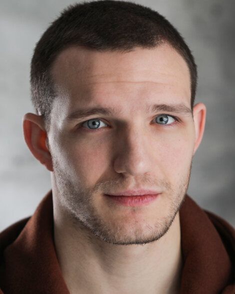 A person with short hair and a neutral expression, wearing a dark shirt against a blurred gray background.