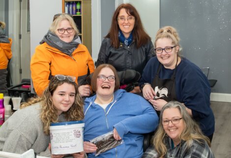 Six women smiling indoors; one holds a bucket labeled "Little Princess Trust," another holds hair extensions. They are casually dressed in a hair salon setting.