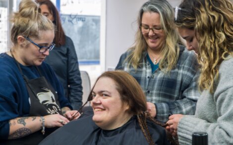 A woman with long hair sits in a salon chair, smiling, as two hairstylists braid her hair while another woman observes.