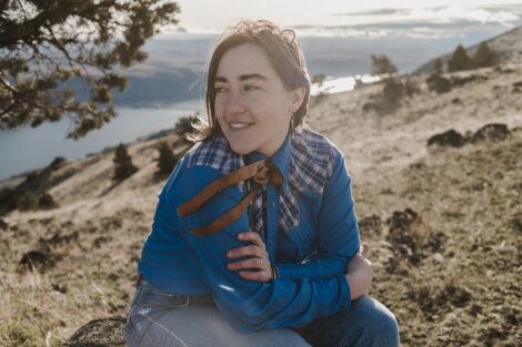 Person in a blue shirt sitting on a hillside, with a scenic view of a river and hills in the background.