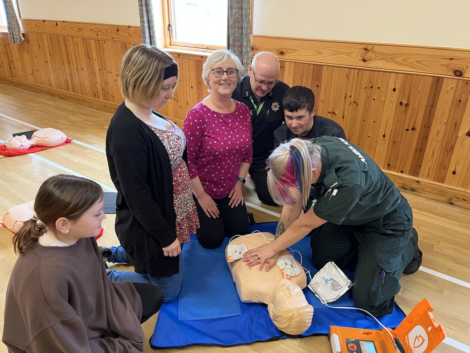 A group of people participates in a CPR training session, observing as an instructor demonstrates on a mannequin.