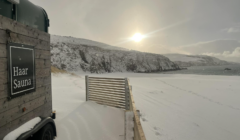 A sauna wagon labeled "Haar Sauna" sits by a snowy beach under a cloudy sky, with hills and the sun in the background.