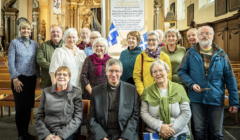 A group of people posing for a photo inside a church, with some holding small flags. A priest is seated in front.