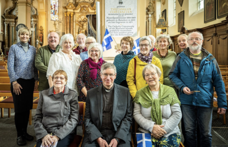 A group of people posing for a photo inside a church, with some holding small flags. A priest is seated in front.
