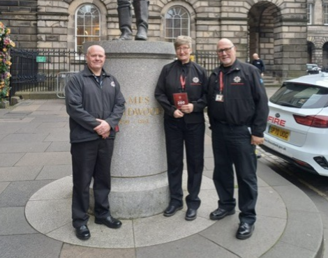 Three uniformed individuals stand in front of a stone monument with engraved text, outdoors on a city street.