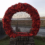 A large wreath made of red poppies is displayed on a stone pedestal in front of a chain-link fence, with a rural landscape in the background.