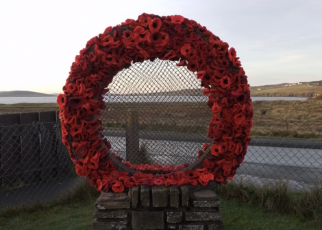 A large wreath made of red poppies is displayed on a stone pedestal in front of a chain-link fence, with a rural landscape in the background.