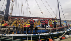 A group of people in waterproof clothing stand on a docked sailing vessel, holding flags. Buoys hang from the boat. It’s overcast with calm waters in the background.