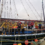 A group of people in waterproof clothing stand on a docked sailing vessel, holding flags. Buoys hang from the boat. It’s overcast with calm waters in the background.