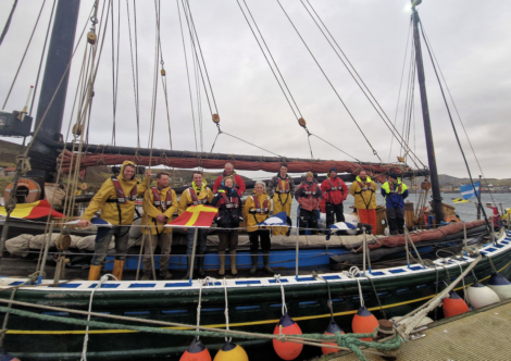 A group of people in waterproof clothing stand on a docked sailing vessel, holding flags. Buoys hang from the boat. It’s overcast with calm waters in the background.