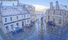 A snow-dusted street scene at Market Cross shows parked cars, historic buildings, and a central monument on a clear day.