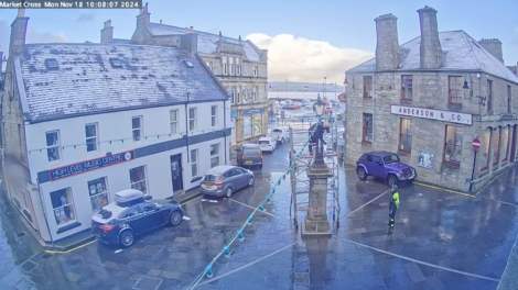 A snow-dusted street scene at Market Cross shows parked cars, historic buildings, and a central monument on a clear day.