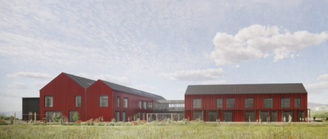 A modern building complex with red exterior, black roofs, and large windows, set in a grassy landscape under a partly cloudy sky.