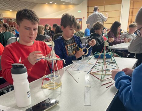 Students sit at tables in a classroom, constructing small structures with straws and clay.