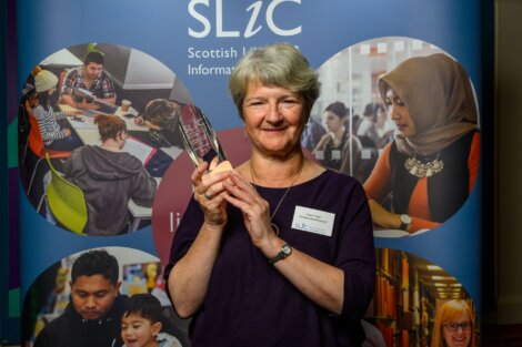 A person holding an award stands in front of a Scottish Library and Information Council backdrop, featuring diverse people engaged in library activities.