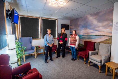 Three people stand in a room with armchairs and a large beach mural on the wall. There are two windows with blinds and a potted plant in the corner.