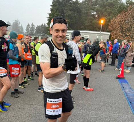 Runner in race attire standing with others at a starting line, wearing a bib labeled "James." Trees and spectators are visible in the background.